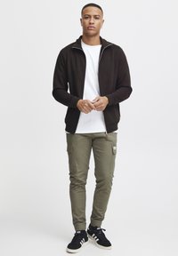Mørkebrun zip-up jakke over en hvit t-skjorte, parret med olivengrønne cargo-bukser. Svarte sneakers med hvite detaljer; minimalistisk design, godt tilpasset utseende.