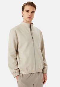 Chaqueta de color beige claro, con cremallera y cuello alto, puños elásticos y bolsillos laterales. Tejido suave con un corte holgado y sin patrones visibles.