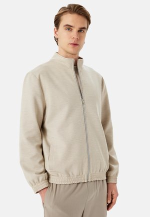 Veste zippée beige clair avec col montant, poignets élastiques et poches latérales. Tissu lisse avec une coupe décontractée et sans motifs visibles.