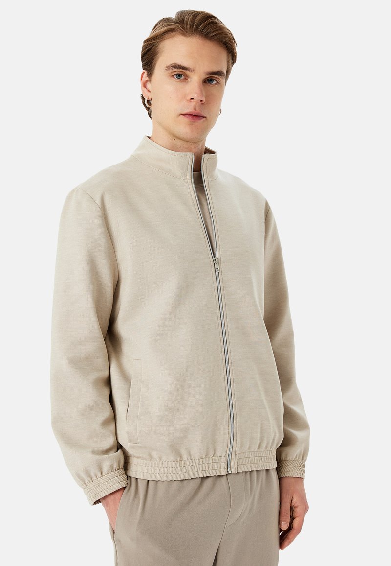 Chaqueta de color beige claro, con cremallera y cuello alto, puños elásticos y bolsillos laterales. Tejido suave con un corte holgado y sin patrones visibles.