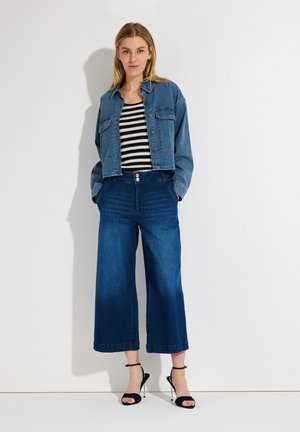 Vrouw die een korte spijkerjack, een mouwloos gestreept topje, wijd uitlopende cropped jeans en zwarte sandalen met hoge hakken draagt, staand tegen een witte muur.