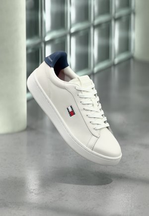 Hvid lædersneaker med marineblå hælflig og rød, hvid og blå logo, der svæver over en poleret betonflade.