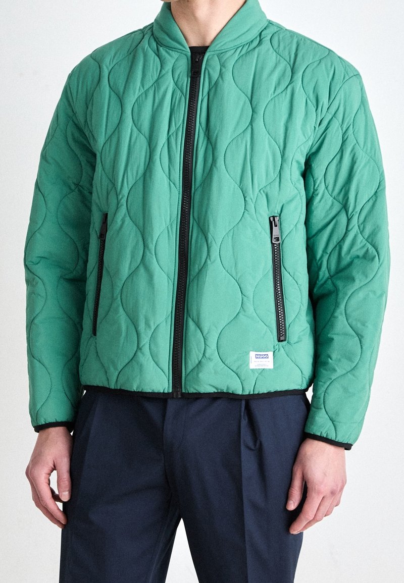 Quilted grøn bomberjakke med et tekstureret bølgemønster, med sorte lynlåse og kant, samt et lille mærke nær kanten.