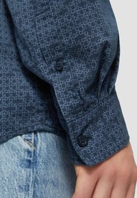 Chemise bleu marine à boutons avec un motif géométrique subtil, en tissu doux, avec des poignets ajustables et des boutons foncés. À associer avec un denim clair.