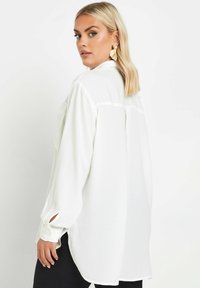 Blusa bianca a maniche lunghe con vestibilità ampia, caratterizzata da dettagli con bottoni sulle maniche e una piega sul retro. Realizzata in tessuto testurizzato.
