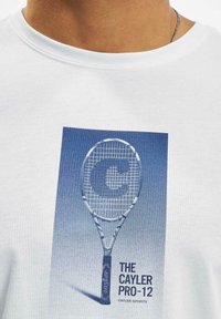 Valge T-särk, millel on sinistes toonides tennisereketi graafika, kus on selgelt näha tekst "THE CAYLER PRO-12" ja "CAYLER SPORTS".