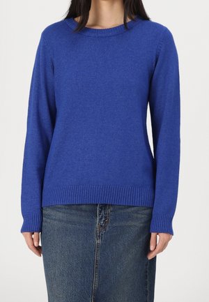 Femme portant un pull bleu roi à manches longues et col rond, associé à une jupe en denim de lavage moyen, debout devant un fond uni.