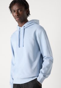 Sudadera con capucha de color azul claro, hecha de un tejido suave, que presenta un bolsillo tipo canguro en la parte frontal, puños acanalados y una marca mínima en el pecho.