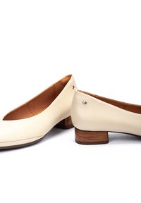 Bailarinas de piel crema con punta redonda, tacón bajo de madera y un sutil acento dorado en el tacón. Textura suave, diseño minimalista.