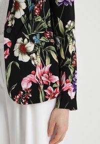 Blazer met bloemenpatroon in zwarte stof. Kenmerkt zich door grote roze, witte en blauwe bloemen met groene bladeren. Inclusief een voorzak en een enkele knoop.