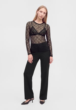 Femme portant un haut en dentelle noire à manches longues avec un soutien-gorge noir en dessous, un pantalon noir à jambes larges et des talons noirs pointus, debout devant un fond uni.