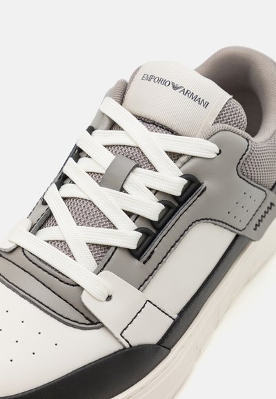 Grijze en witte sneaker met een bovenwerk van leer en mesh, voorzien van witte veters, een zwarte neus en branding op de tong.