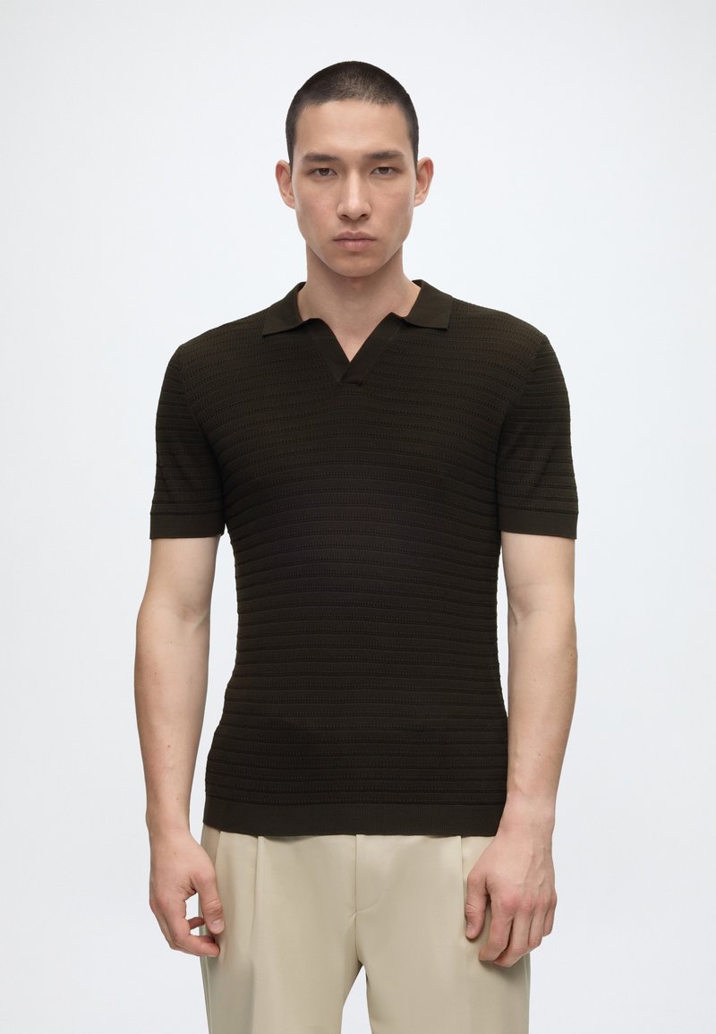 Homme portant un polo texturé à manches courtes de couleur foncée et un pantalon beige, debout devant un fond blanc uni.