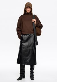 Brązowa, oversize'owa bluza, czarna spódnica z ekologicznej skóry, wysokie czarne botki na obcasie, brązowa czapka z logo, trzymająca brązową torbę z paskiem.