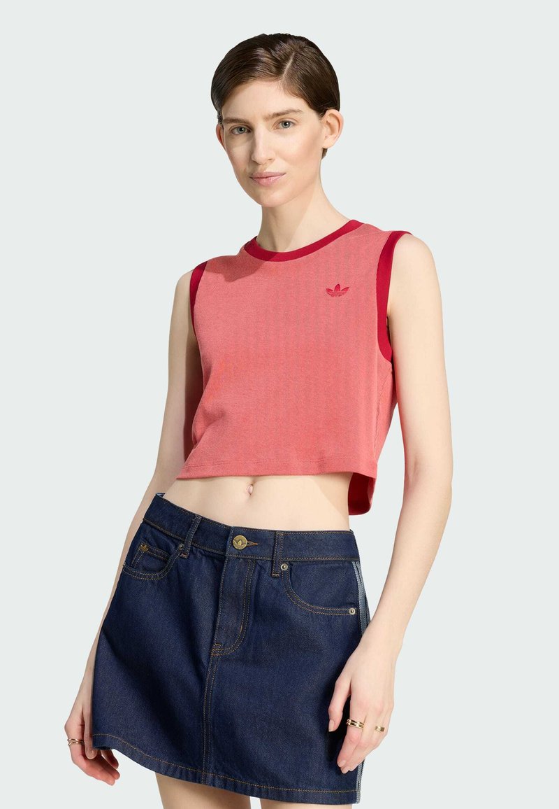 Ung kvinde iført en ærmeløs rød crop top og mørkeblå denim miniskørt poserende med hånden hvilende nær hoften mod hvid baggrund.