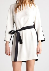 Robe blanche avec des manches trois-quarts et une ceinture noire. Coupe droite et accents latéraux noirs. Texture de tissu lisse.