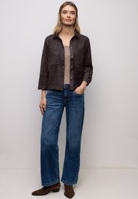 Chaqueta de ante marrón con cuello, mangas tres cuartos y dos bolsillos delanteros, combinada con vaqueros de mezclilla azul de pierna ancha y botines marrones.