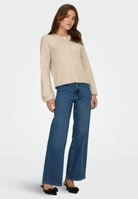 Beige gestrickte Strickjacke mit rundem Ausschnitt und Perlenknöpfen, kombiniert mit weit geschnittenen blauen Jeans und schwarzen Mules mit Schleifenakzent.