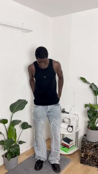 Camisola preta sem mangas, jeans desgastados azul claro com bordas desfiadas, sapatos de couro pretos, em pé entre plantas de interior.