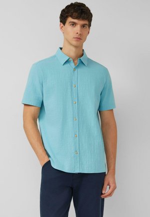 Junger Mann, der ein hellblaues kurzärmeliges Hemd und dunkelblaue Hose trägt, steht mit einer Hand in der Tasche.