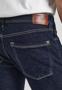 Acercamiento de un bolsillo trasero de unos jeans de mezclilla azul oscuro con costuras amarillas y una etiqueta de marca de cuero marrón encima del bolsillo, en una persona que lleva una camisa blanca.