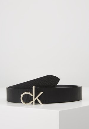 Ceinture en cuir texturé noir avec boucle logo "ck" en argent, présentée enroulée sur une surface blanche avec un fond gris uni.