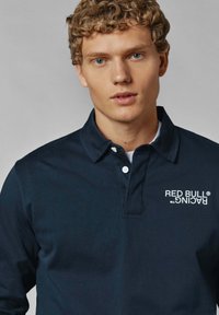 Temno modra polo majica z zapenjanjem na tri gumbe in belim logotipom "Red Bull Racing" na levem prsnem delu. Mehka bombažna tkanina.