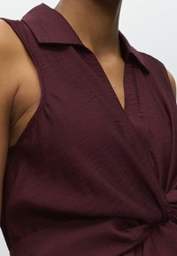 Gros plan d'une personne portant un haut sans manches bordeaux foncé avec un col et un détail froncé à la taille.