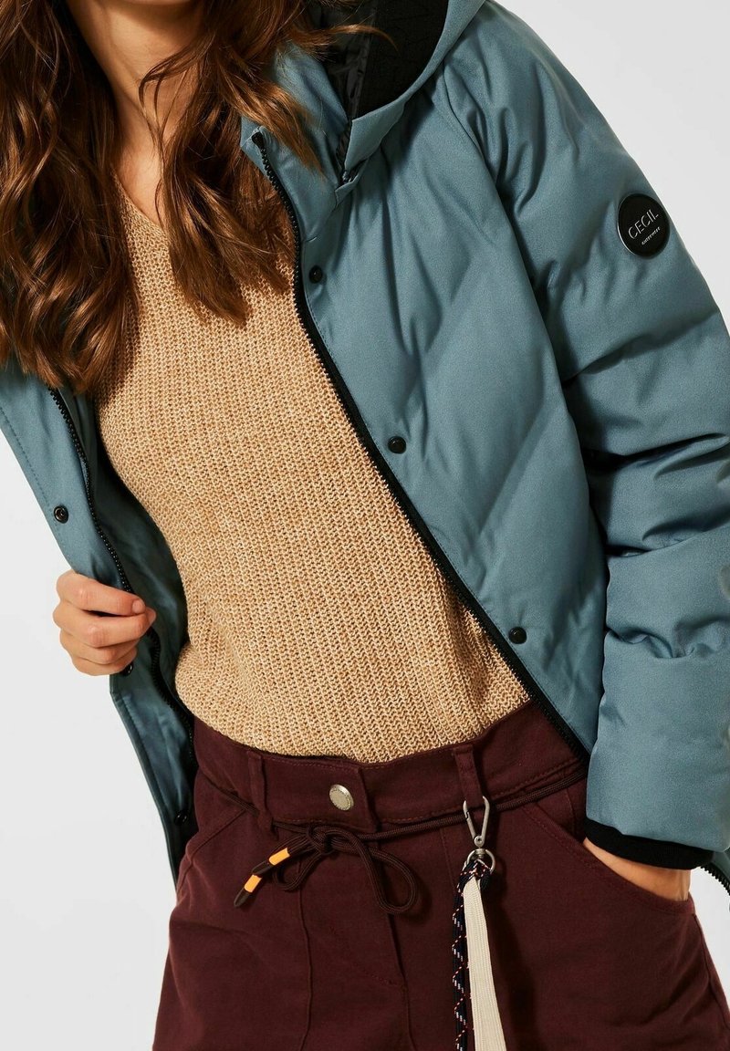 Femme portant une doudoune turquoise par-dessus un pull beige en tricot et un pantalon bordeaux foncé avec un cordon de serrage et un porte-clés suspendu.