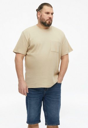 Homme avec barbe et cheveux attachés, portant une chemise beige à manches courtes et un short en jean bleu, debout avec une main dans la poche.