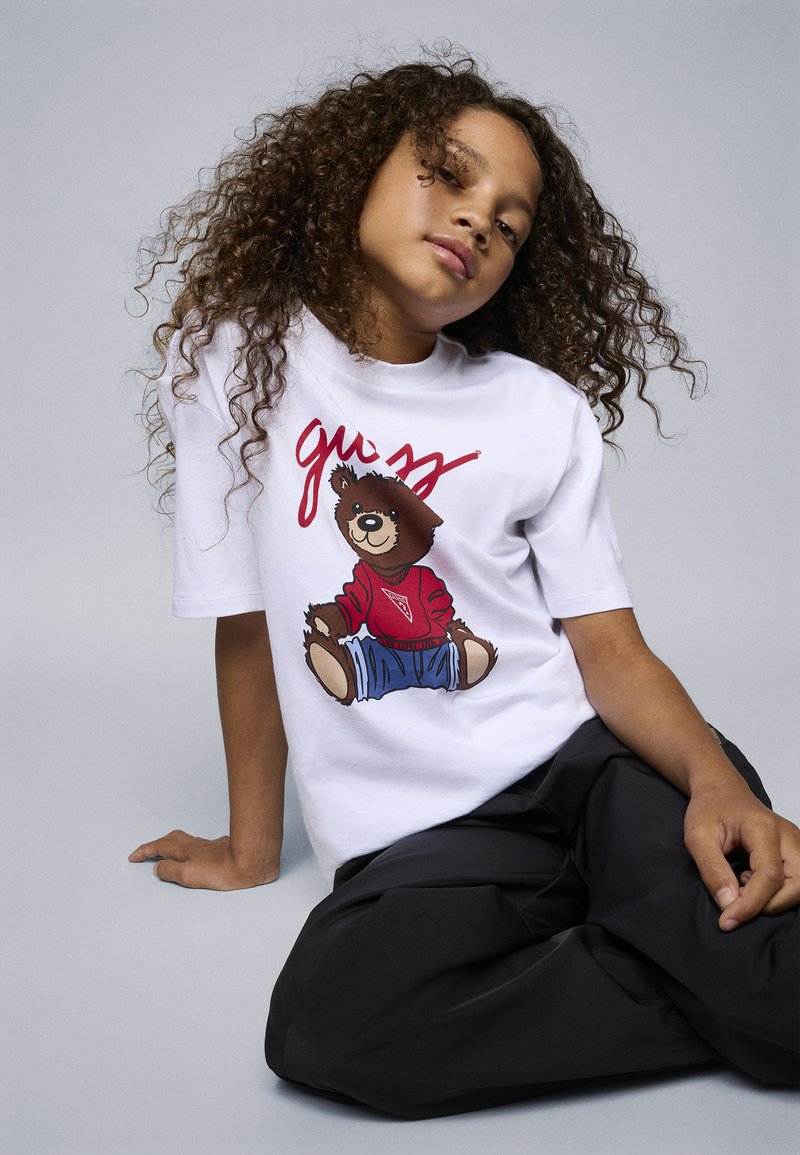 Vit t-shirt med korta ärmar som visar en tecknad björn i en röd tröja och blå jeans, med texten "guess" i rött ovanför. Lossa svarta byxor.