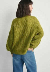 Gruby zielony sweter z dzianiny o wzorze warkocza, szerokimi rękawami i lekko krótkim kroju, zestawiony z luźnymi niebieskimi dżinsami.