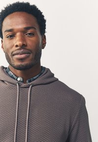 Felpa con cappuccio marrone imbottita con coulisse, indossata sopra una camicia a quadri blu. Primo piano che mostra dettagli di tessitura e cuciture.