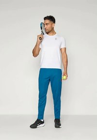 T-shirt branca de mangas curtas, calças de atletismo azuis, ténis pretos. A segurar uma raquete de ténis numa mão e uma bola de ténis na outra.