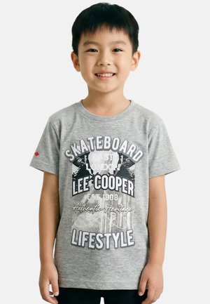 Garçon souriant portant un T-shirt gris à thème skateboard avec le texte "Lee Cooper Lifestyle", debout devant un fond clair uni.