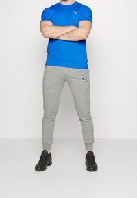 Modrá atletická tričko s krátkým rukávem, šedé tepláky s manžetami na kotnících a černé tenisky. Na tričku a kalhotách je logo Puma.