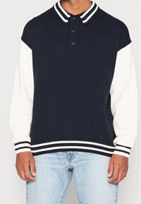 Suéter azul marino con mangas blancas, textura acanalada, cuello a rayas y puños. Presenta una tapeta con tres botones.