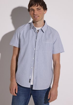 Homme portant une chemise à manches courtes rayée bleu et blanc à boutons par-dessus un t-shirt blanc et un jean bleu, debout devant un fond clair uni.