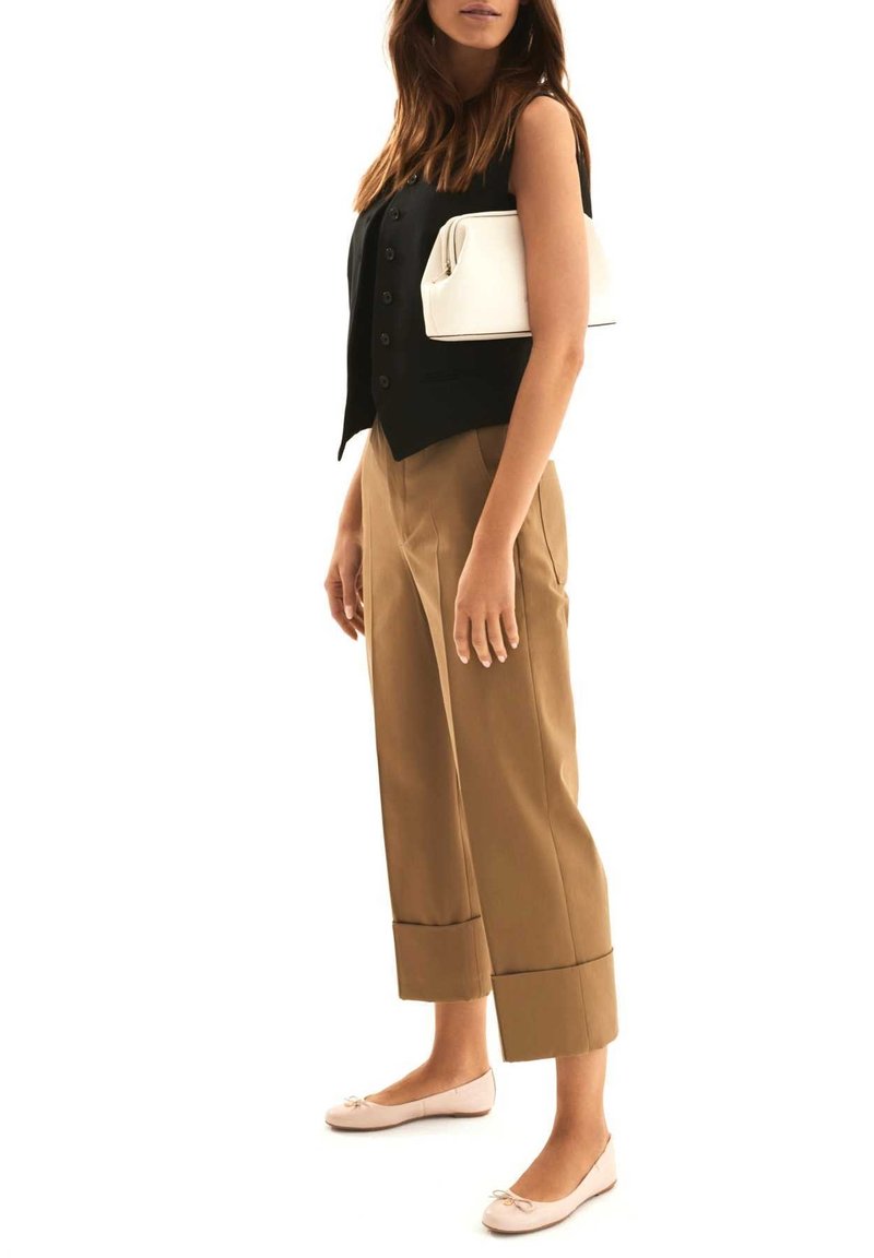 Femme portant un gilet noir sans manches à boutons, un pantalon court couleur beige, des ballerines rose pâle et tenant une pochette blanche sous le bras.