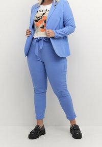 Costume bleu clair avec un blazer cintré et un pantalon fuselé. La tenue est associée à un t-shirt blanc à imprimés orange et des chaussures noires.