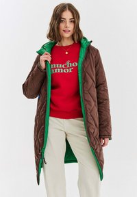 Prešívaná hnedá a zelená bunda s kapucňou, so zapínaním na zips a dvoma bočnými vreckami. Nosená cez červený sveter so zeleným textom.