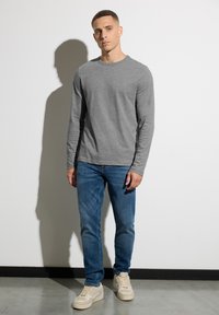 Graues Langarmshirt mit Rundhalsausschnitt, kombiniert mit blauen Jeans und beigen Sneakers. Glatte Textur mit minimalen Details.