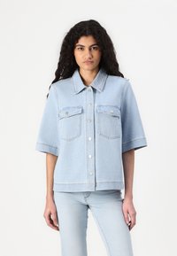 Casaco de ganga azul claro com mangas curtas, dois bolsos frontais, botões prateados e colarinho de camisa; ajuste ligeiramente solto com detalhes costurados.