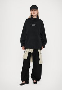 Black oversized fleece sweatshirt with a high collar and logo. Paired with loose black pants and a beige shirt tied at the waist. Black cap and flats.