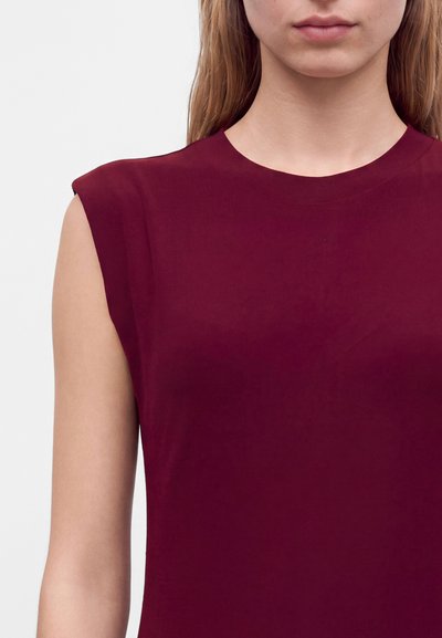 Gros plan d'une femme portant un haut sans manches rouge foncé à encolure ronde sur un fond clair uni.