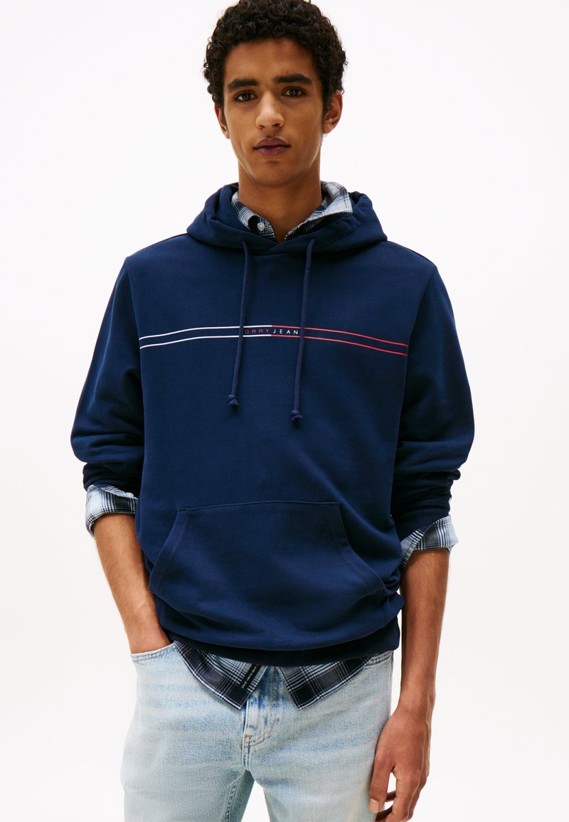 Marineblå hettegenser med hvite og røde horisontale striper, kengurulomme og snorer. Worn over en blå rutete skjorte og lyse jeans.