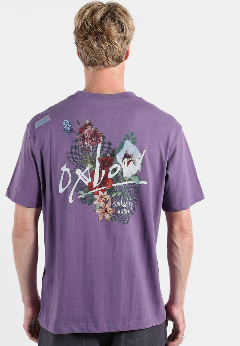 T-shirt en coton violet avec un motif floral au dos, comprenant un mélange de plantes et un texte en gras, manches courtes et coupe décontractée.