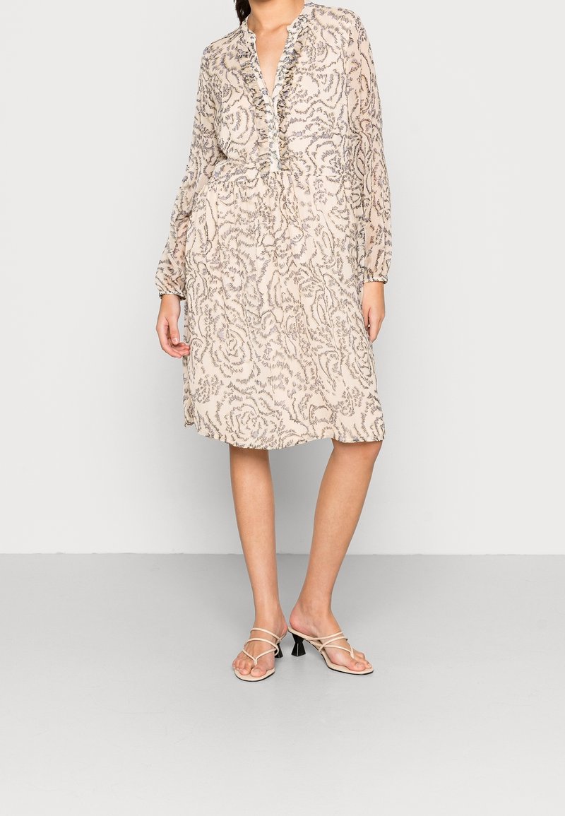 Světle béžové šaty s květinovým vzorem, volány kolem výstřihu, dlouhé rukávy a projmutý pas. Spárováno s sandály na podpatku se zavazováním.