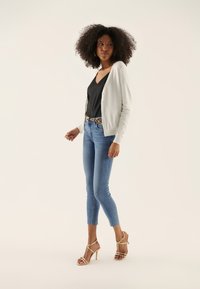 White cardigan, black v-neck top, light blue skinny jeans, and beige high-heeled sandals. Model stands in a relaxed pose against a light background.