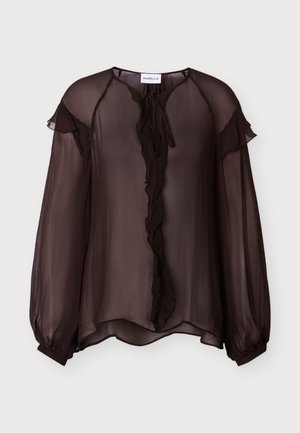 Femme portant un chemisier marron transparent à manches longues avec des volants et un pantalon marron à rayures larges, debout sur un fond blanc uni.